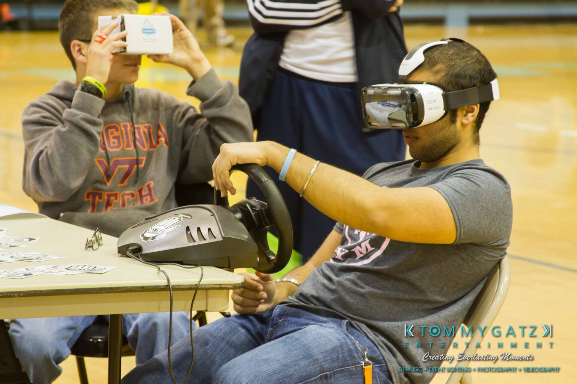 Ford-Driving-Skills-For-Life-15 - TOMMY GATZ ENTERTAINMENT Two people using virtual reality headsets. One has a cardboard VR viewer, the other operates a steering wheel controller. They're seated at a table in a gymnasium.