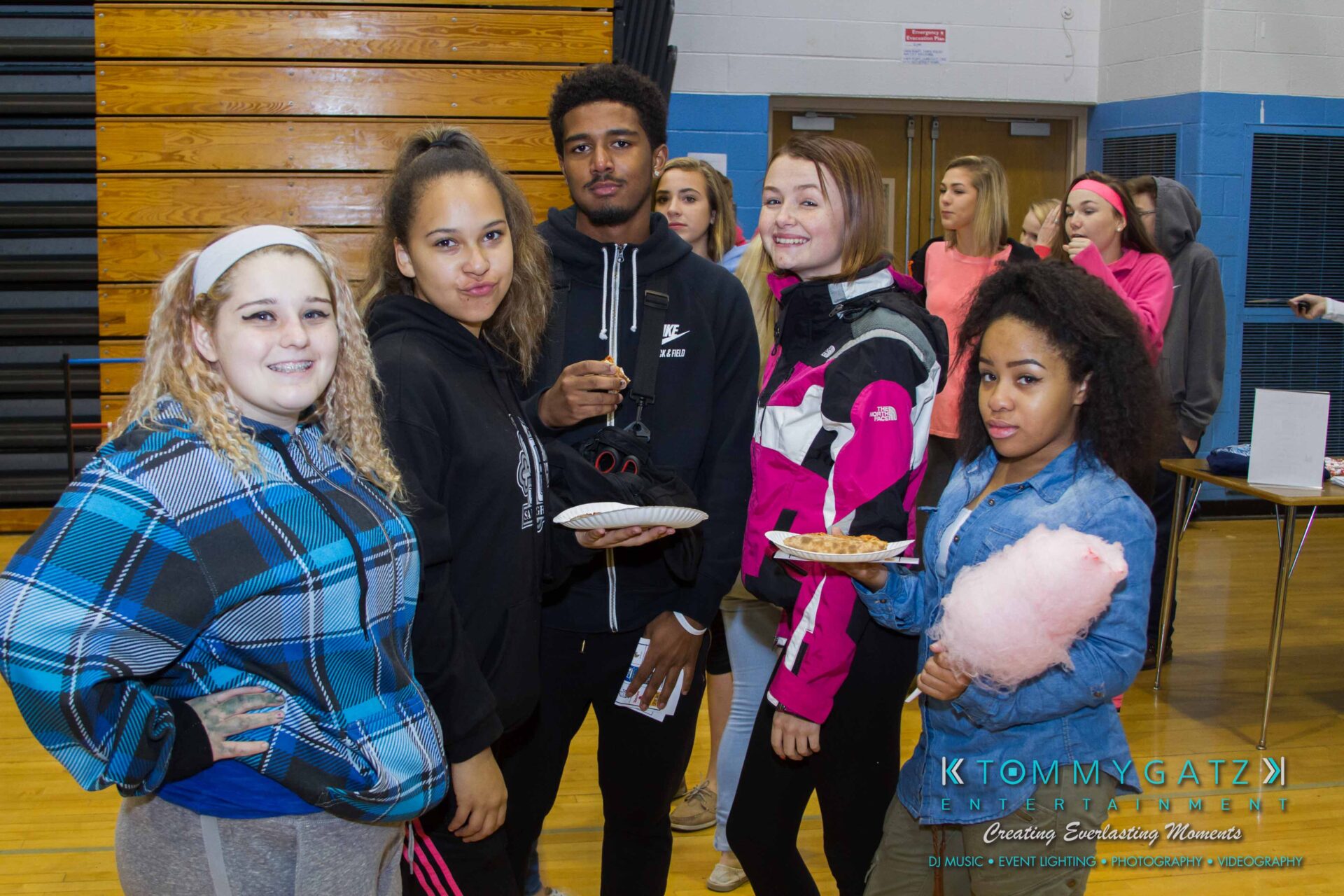 Ford-Driving-Skills-For-Life-21 - TOMMY GATZ ENTERTAINMENT A group of five young people stand indoors. Two hold plates with food, and one holds cotton candy. They are casually dressed in a gymnasium setting.