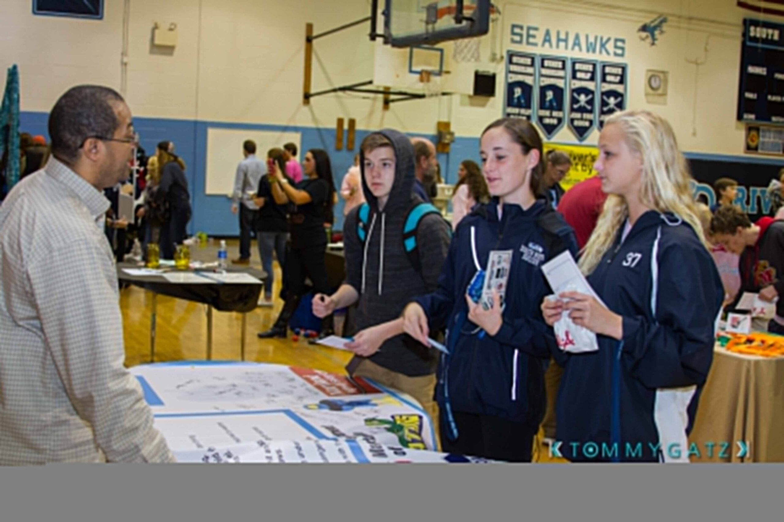 Ford-Driving-Skills-For-Life-26 - TOMMY GATZ ENTERTAINMENT Three people stand in a gymnasium, talking to a man behind a table with brochures and informational materials. Other attendees and tables are visible in the background.