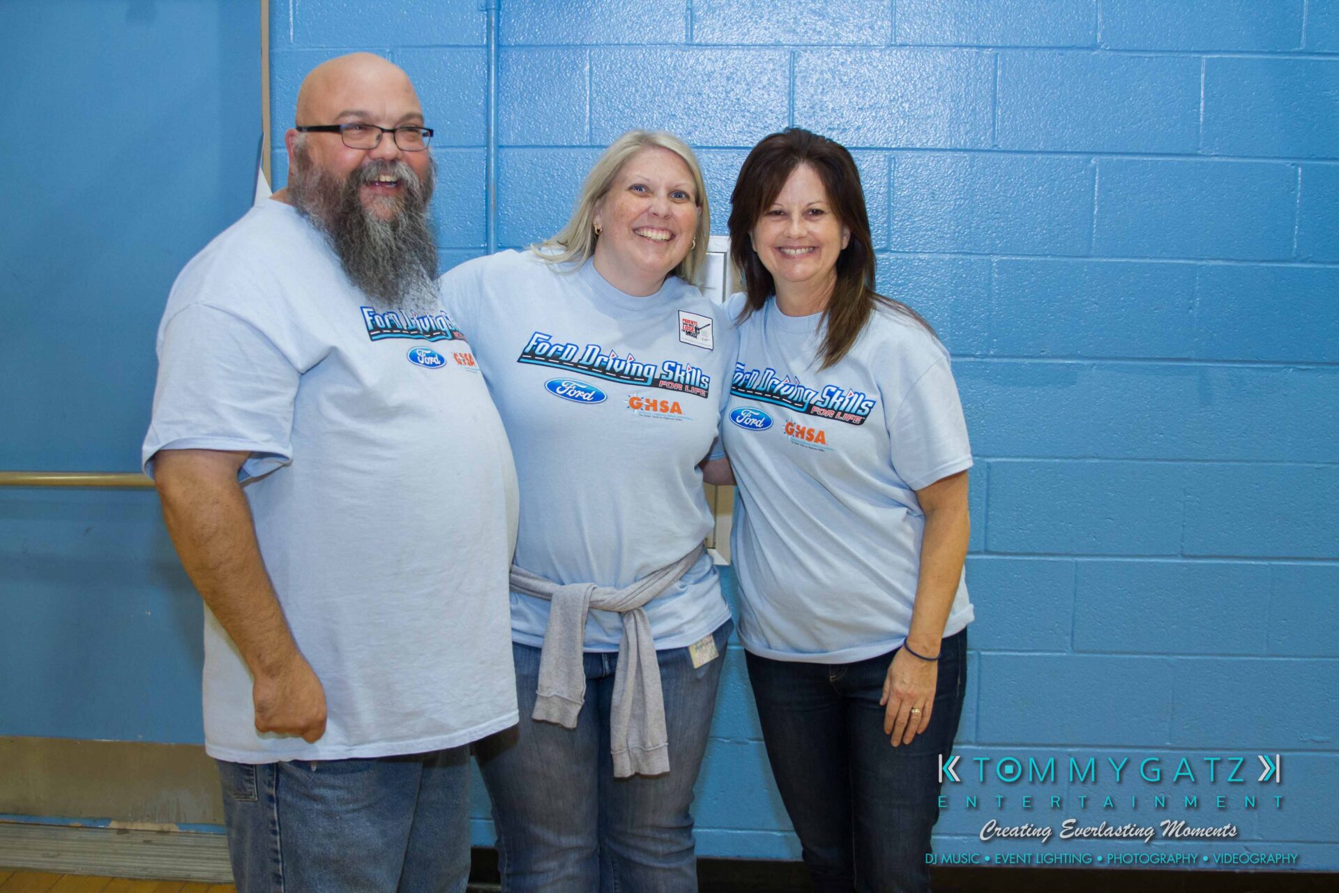 Ford-Driving-Skills-For-Life-32 - TOMMY GATZ ENTERTAINMENT Three people stand smiling, wearing matching light blue shirts with logos. They are indoors against a blue wall.