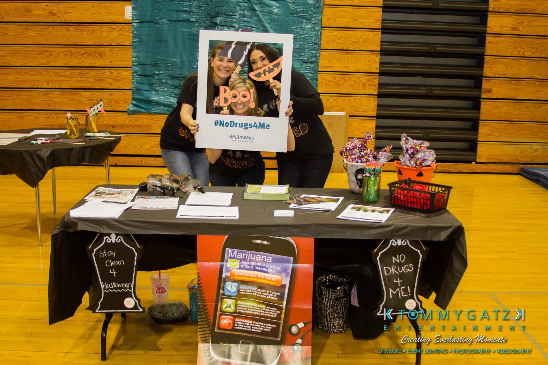 Ford-Driving-Skills-For-Life-33 - TOMMY GATZ ENTERTAINMENT Two people holding a "#NoDrugs4Me" sign behind a table with educational materials and Halloween-themed decorations in a gymnasium.