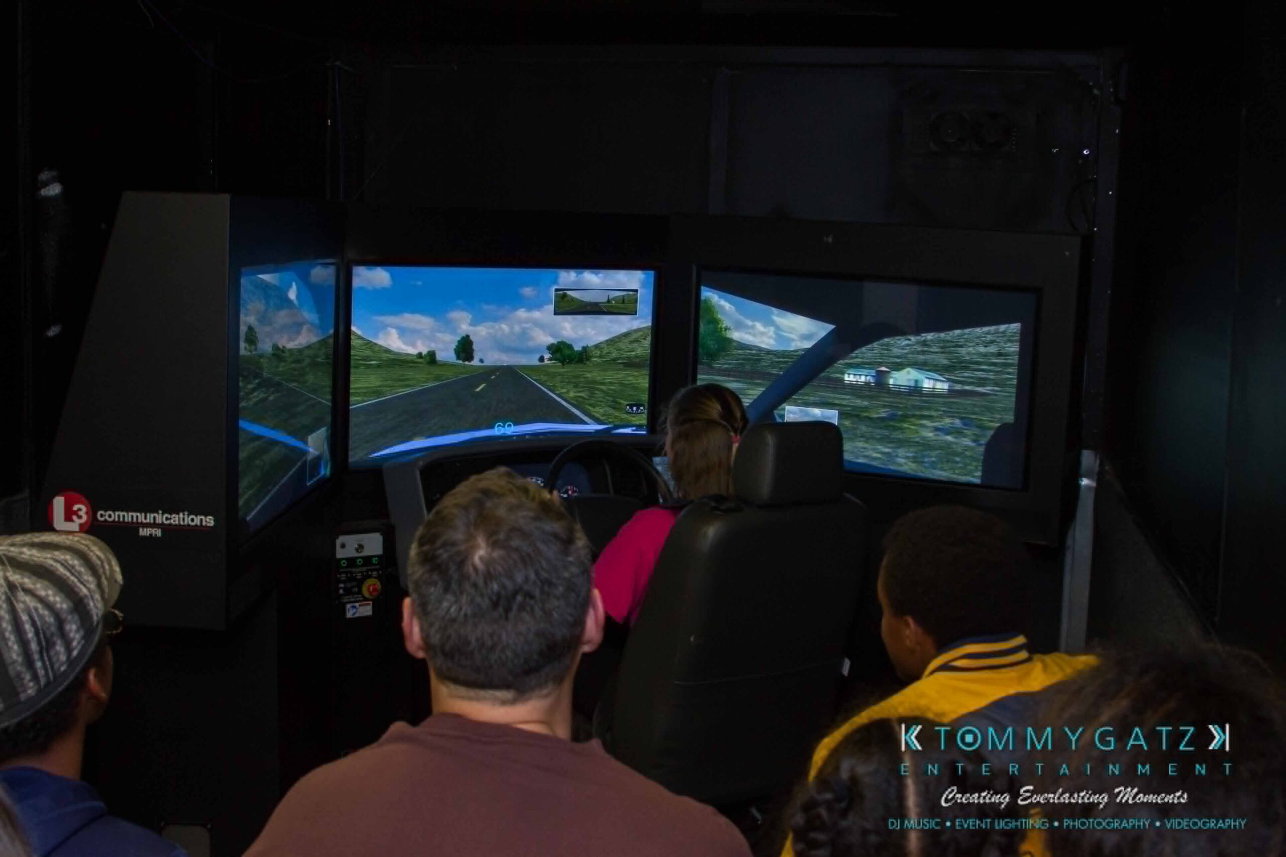 Ford-Driving-Skills-For-Life-36 - TOMMY GATZ ENTERTAINMENT A person drives a car simulation setup with three screens displaying a racing game. Several people watch from behind.