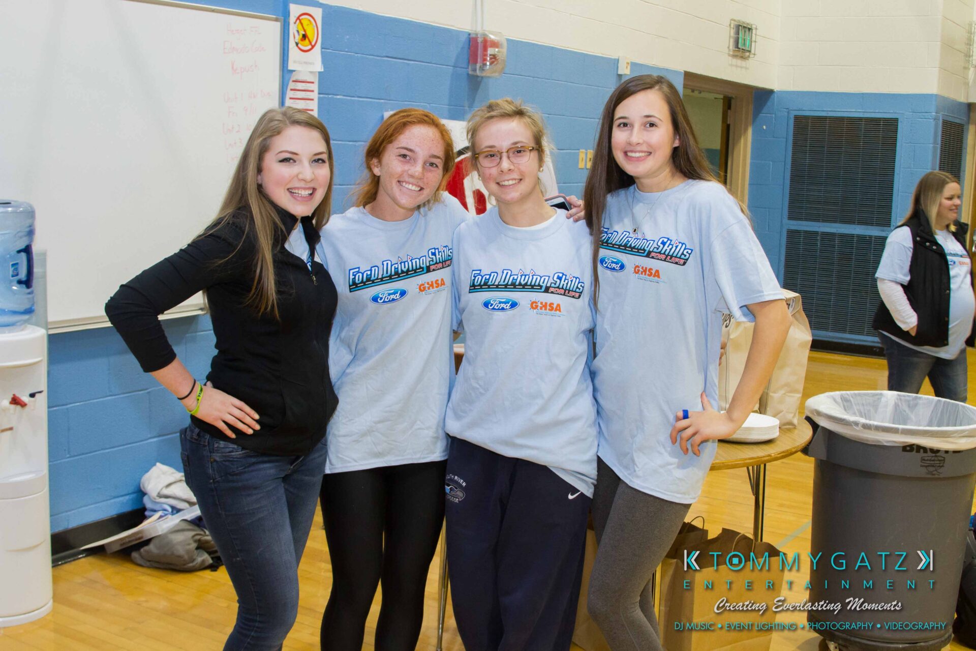 Ford-Driving-Skills-For-Life-38 - TOMMY GATZ ENTERTAINMENT Four people smiling and posing together in a school gymnasium next to a whiteboard and recycling bin.