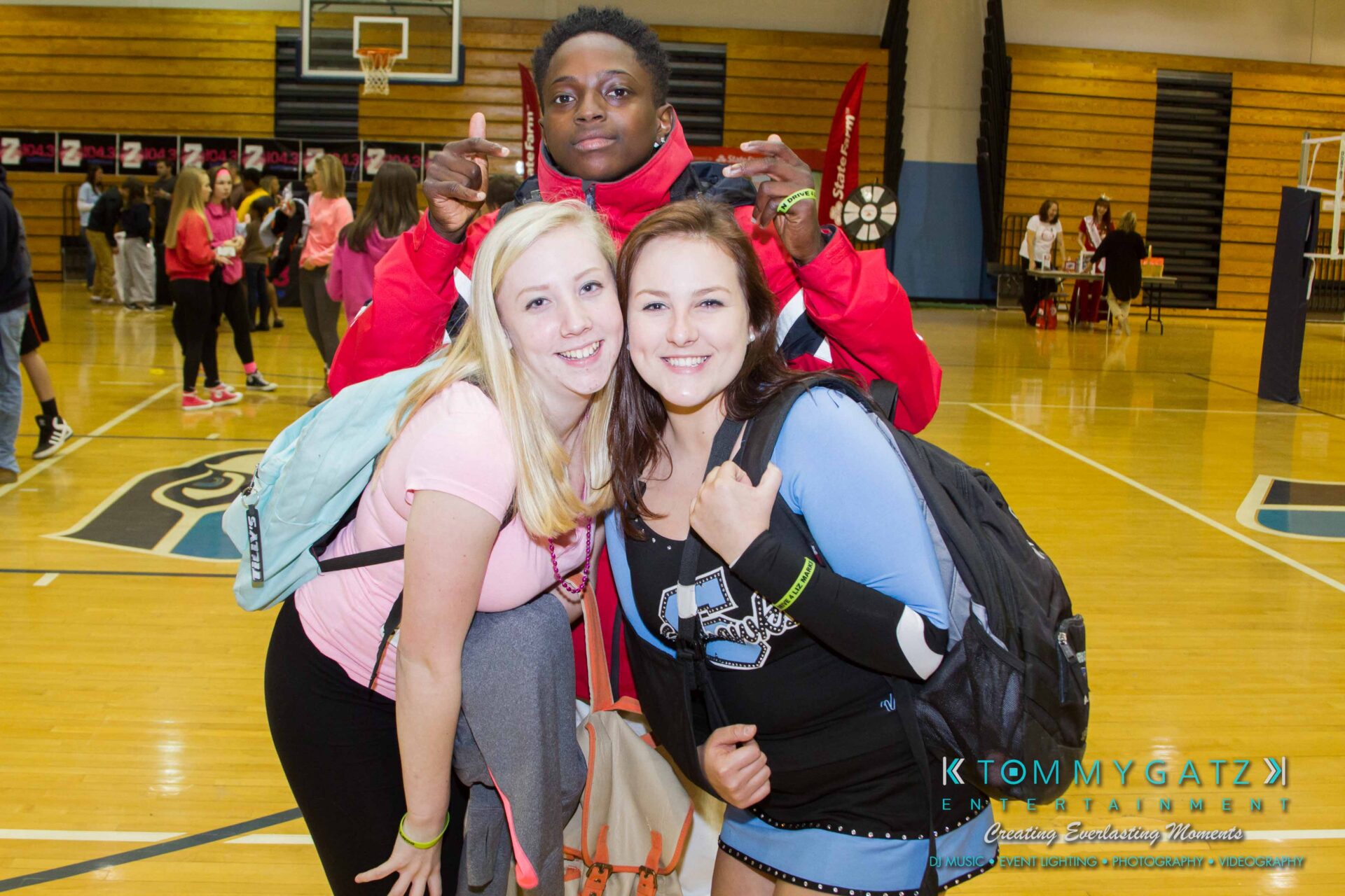 Ford-Driving-Skills-For-Life-39 - TOMMY GATZ ENTERTAINMENT Three people smiling in a gymnasium with others in the background. Two women are in the front with backpacks, and a man stands behind them.