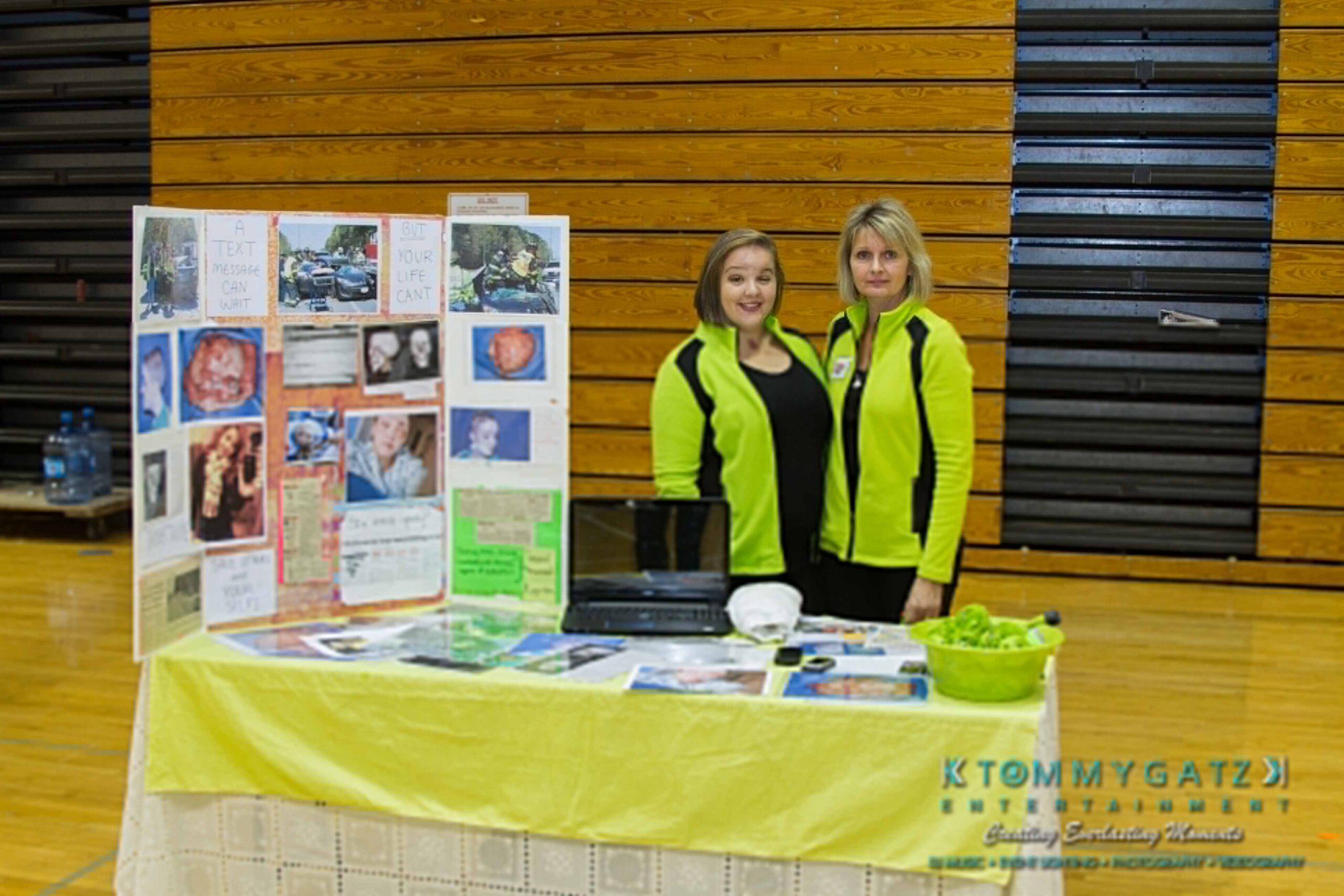 Ford-Driving-Skills-For-Life-4 - TOMMY GATZ ENTERTAINMENT Two people stand behind a display table with a colorful project board and items, in a gymnasium. They are wearing matching green and black outfits.
