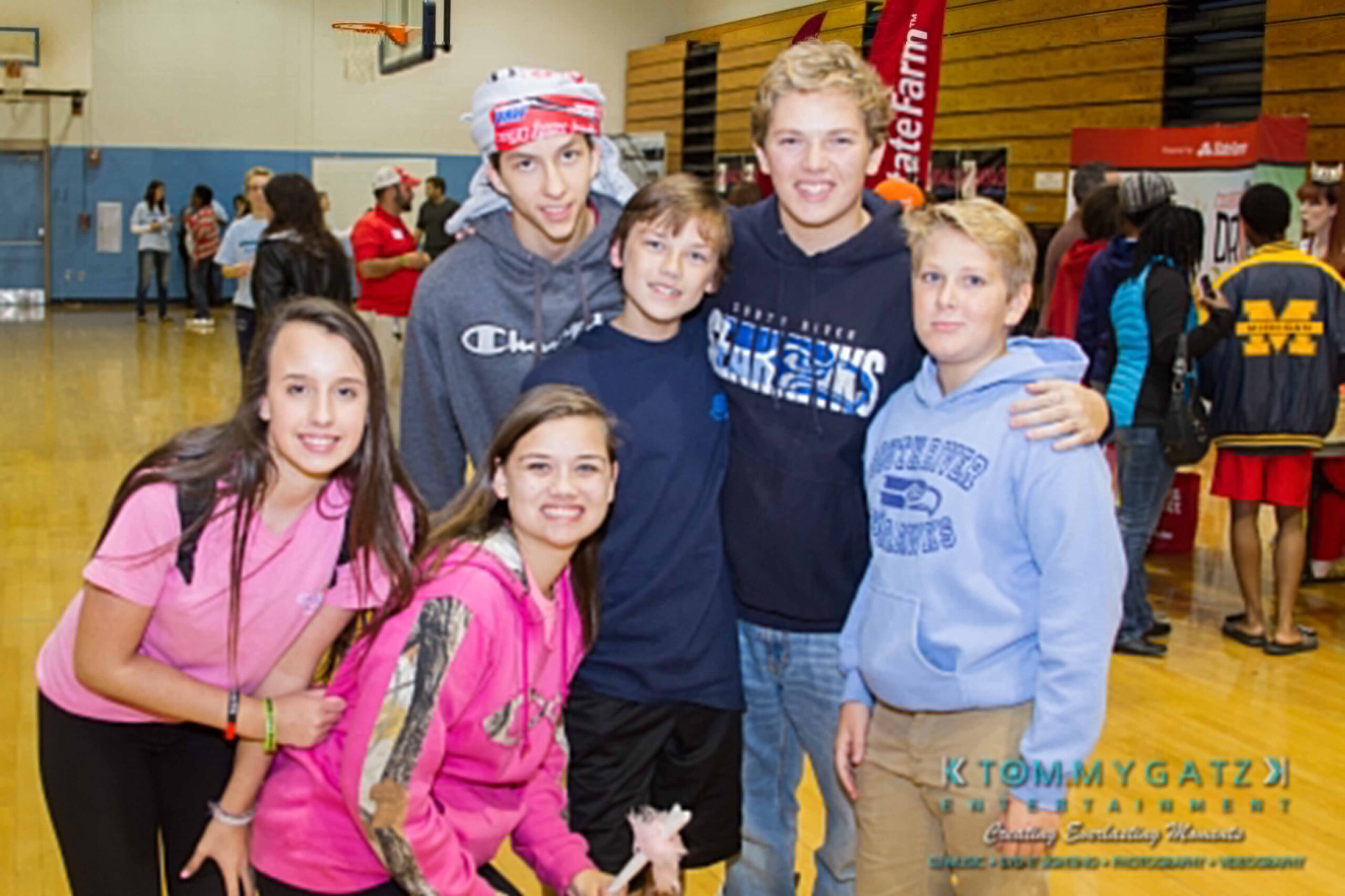 Ford-Driving-Skills-For-Life-41 - TOMMY GATZ ENTERTAINMENT A group of six smiling teenagers pose together in a gymnasium, wearing casual clothing. Several booths and people are visible in the background.