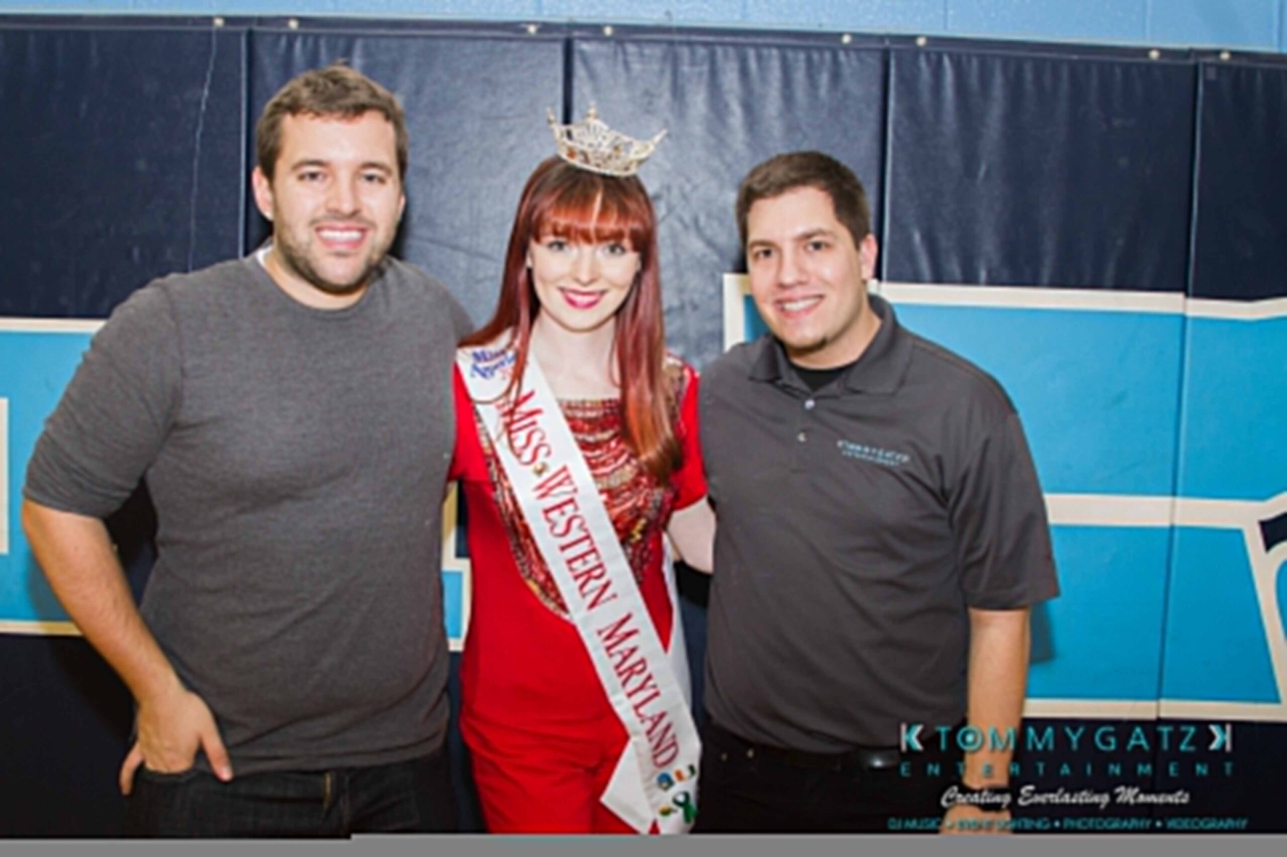 Ford-Driving-Skills-For-Life-47 - TOMMY GATZ ENTERTAINMENT Three people are posing together. The woman in the center wears a crown and a "Miss Western Maryland" sash. Two men stand on either side, one wearing a gray shirt, the other a branded polo.
