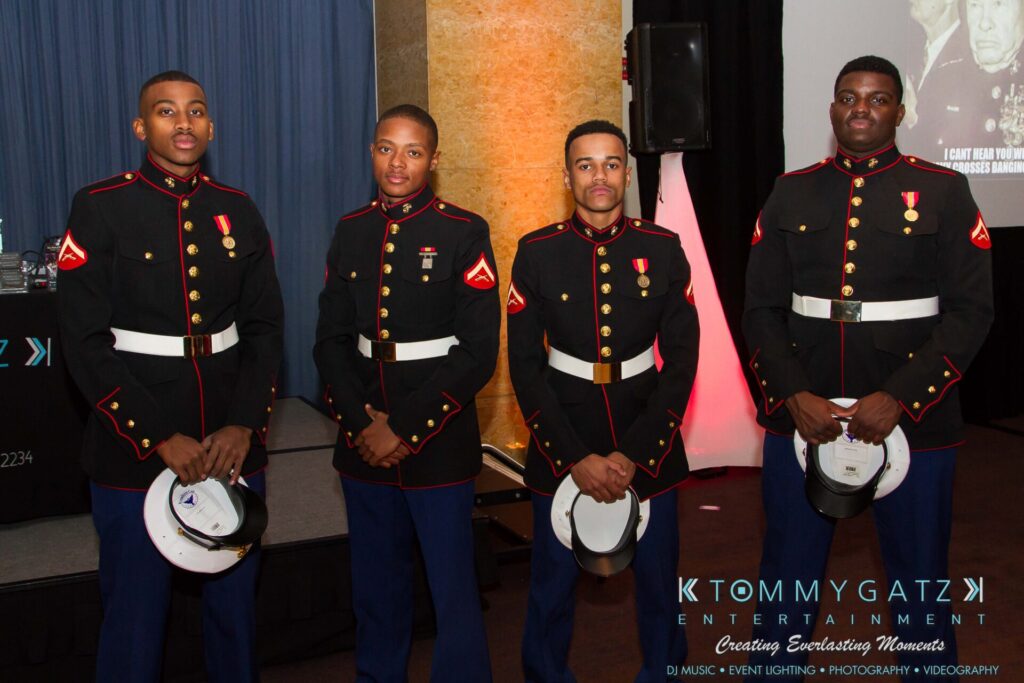 Marine-Lo-Res-10 - TOMMY GATZ ENTERTAINMENT Four Marines in dress uniforms stand in a line, holding white caps. A wall displays a partial image and signage for Tommy Gatz Entertainment in the background.