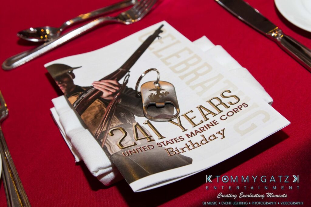 Marine-Lo-Res - TOMMY GATZ ENTERTAINMENT Close-up of a Marine Corps birthday program on a table with a napkin, silverware, and a keychain with a dog tag design.
