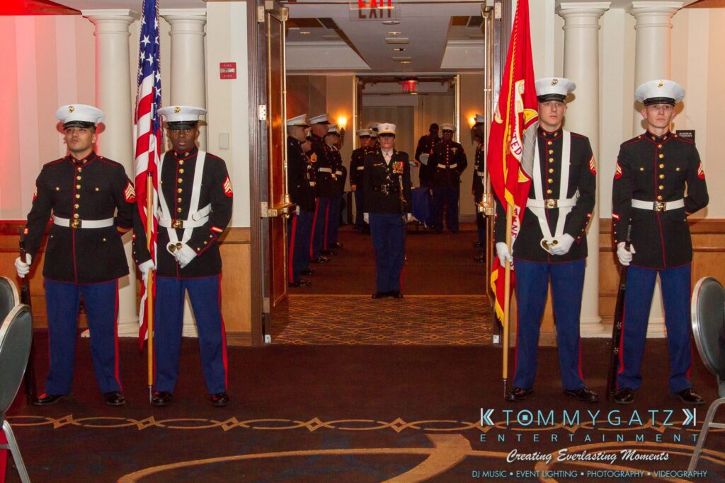 Marine-Lo-Res-11 - TOMMY GATZ ENTERTAINMENT Marines in formal dress uniforms stand in formation with flags, as an officer approaches from a hallway.