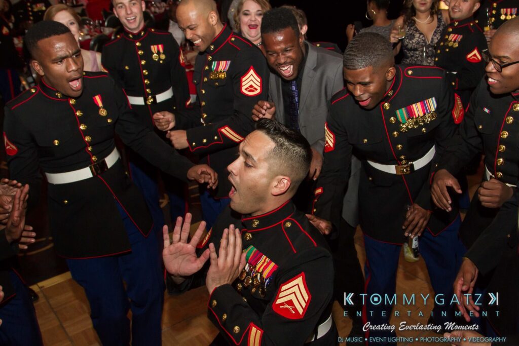 Marine-Lo-Res-118 - TOMMY GATZ ENTERTAINMENT A group of Marines and a man in a suit joyfully celebrate and dance at an event.