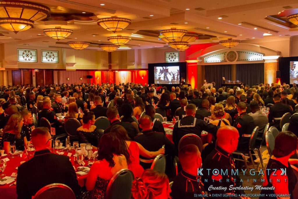 Marine-Lo-Res-12 - TOMMY GATZ ENTERTAINMENT A large, formal event with seated guests in a banquet hall. The room is decorated with chandeliers and red lighting. A screen displays an image at the front.
