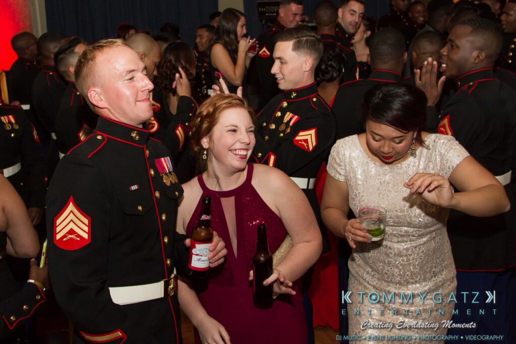 Marine-Lo-Res-129 - TOMMY GATZ ENTERTAINMENT People in formal military attire and evening wear are enjoying a social event, holding drinks and smiling in a crowded room.