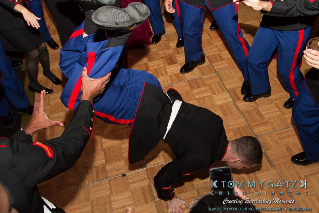 Marine-Lo-Res-131 - TOMMY GATZ ENTERTAINMENT A person in a military uniform performs a breakdance move on a wooden floor while surrounded by others in similar attire.