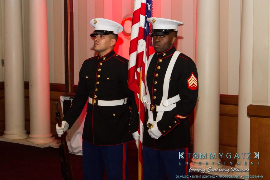 Marine-Lo-Res-15 - TOMMY GATZ ENTERTAINMENT Two uniformed Marines stand at attention holding a flag in a formal indoor setting with white columns.
