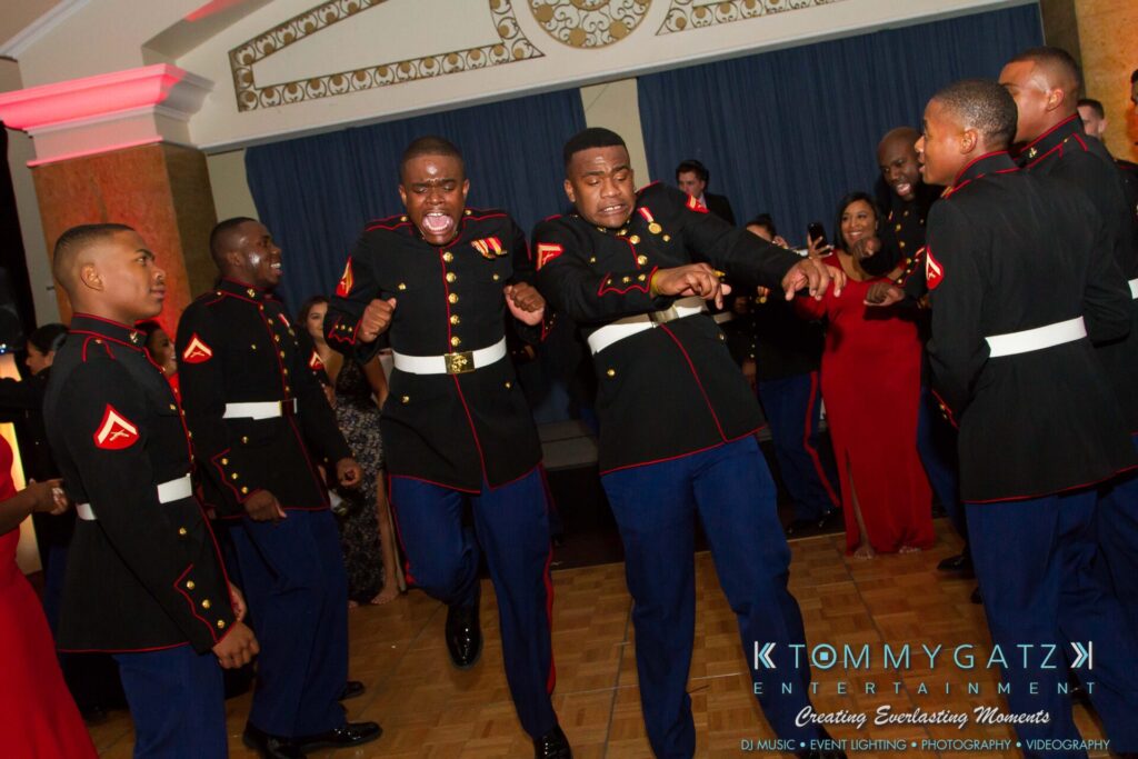 Marine-Lo-Res-166 - TOMMY GATZ ENTERTAINMENT Marines in dress uniforms dance energetically at an event, with people watching and participating in the background.