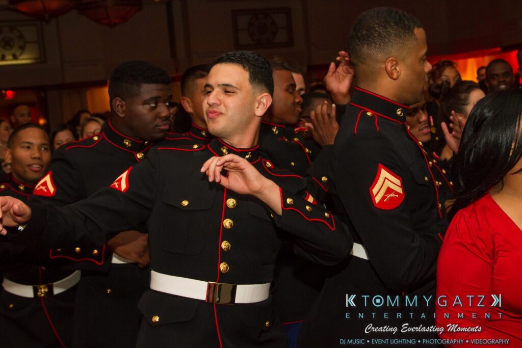 Marine-Lo-Res-180 - TOMMY GATZ ENTERTAINMENT A group of people in formal military attire are gathered indoors, with one person in the foreground dancing.