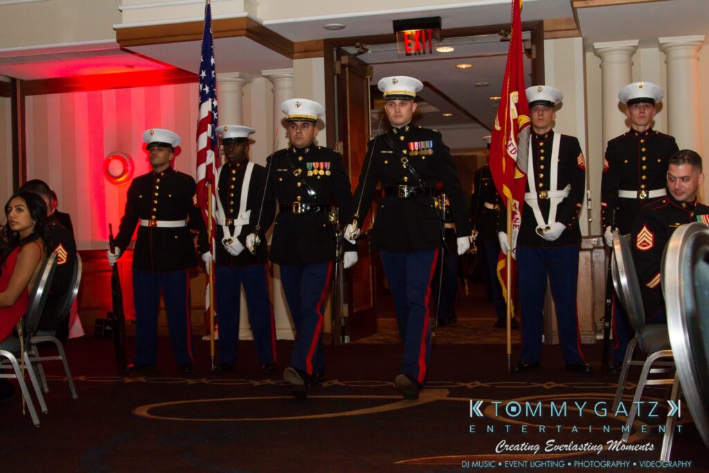 Marine-Lo-Res-19 - TOMMY GATZ ENTERTAINMENT A group of Marines in dress uniforms march through a dimly lit room, flanked by the U.S. flag and Marine Corps flag. People are seated on both sides of the group.