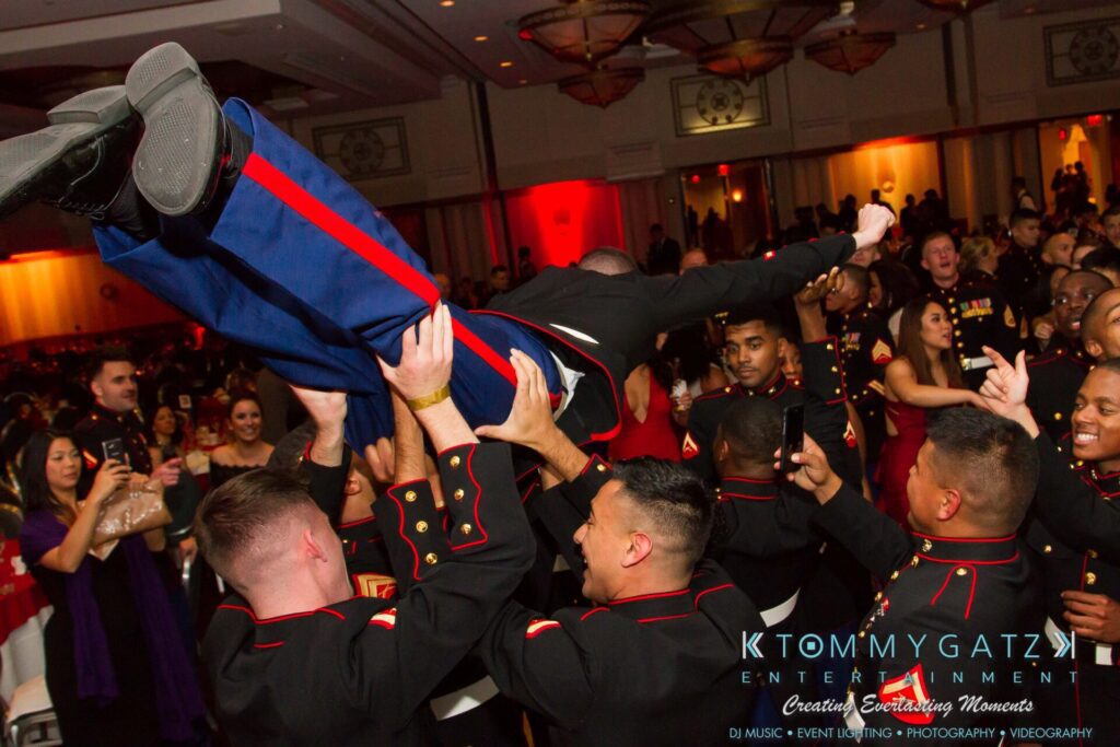 Marine-Lo-Res-192 - TOMMY GATZ ENTERTAINMENT Group of people in formal military uniforms lift a person in the air at a lively indoor event.