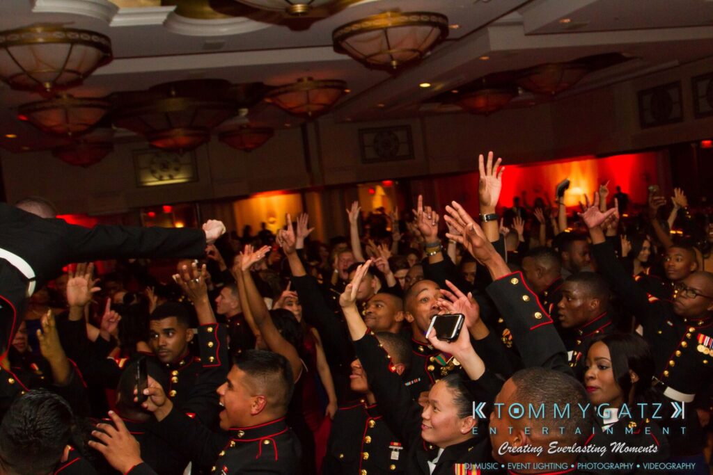 Marine-Lo-Res-193 - TOMMY GATZ ENTERTAINMENT A group of people in uniform cheer and reach towards the stage at an indoor event with dim lighting.