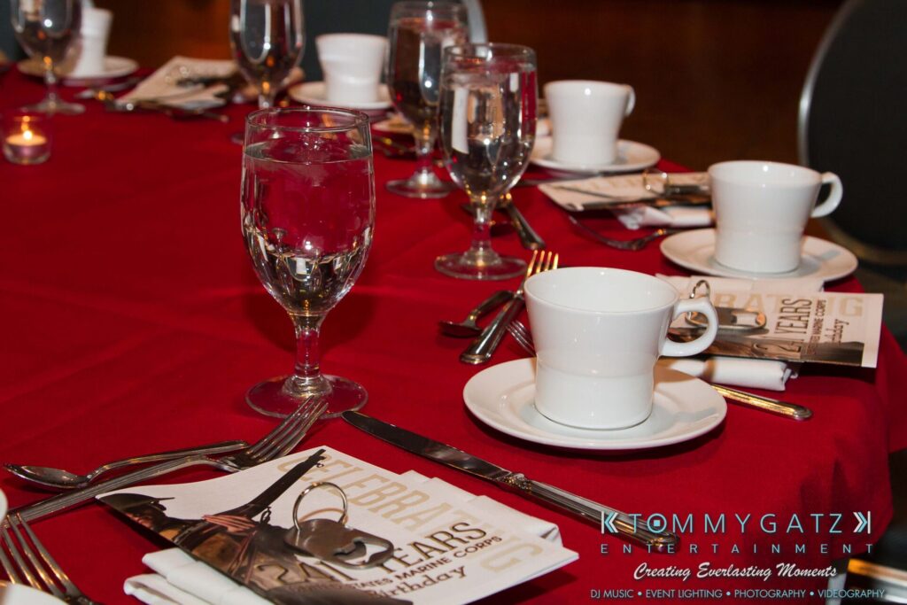Marine-Lo-Res-2 - TOMMY GATZ ENTERTAINMENT A red tablecloth with neatly arranged dinnerware, including cups, saucers, and glasses. A folded napkin and booklets are placed by each setting.