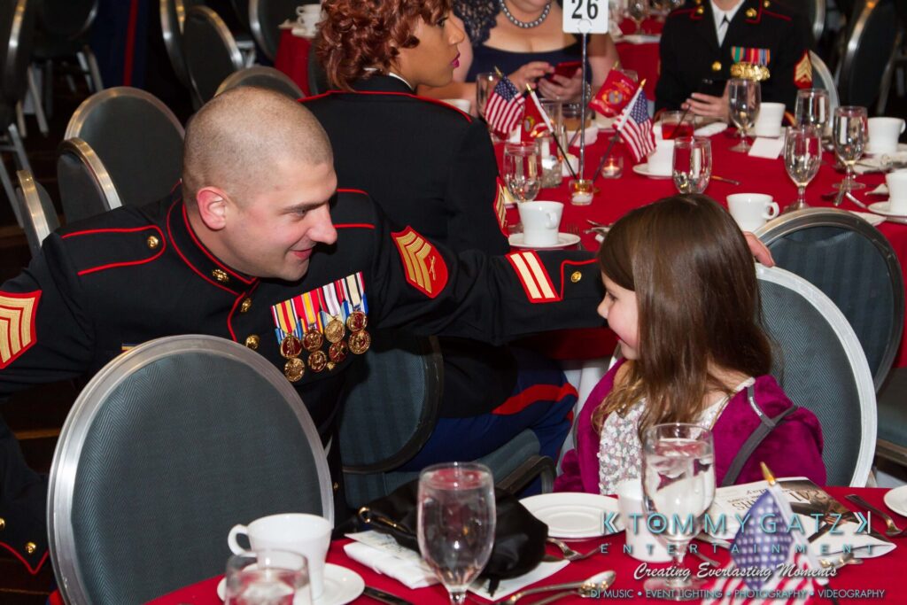 Marine-Lo-Res-3 - TOMMY GATZ ENTERTAINMENT A Marine in dress uniform talks to a smiling young girl at a formal event. They are seated at a table with red tablecloths, decorated with small American flags.