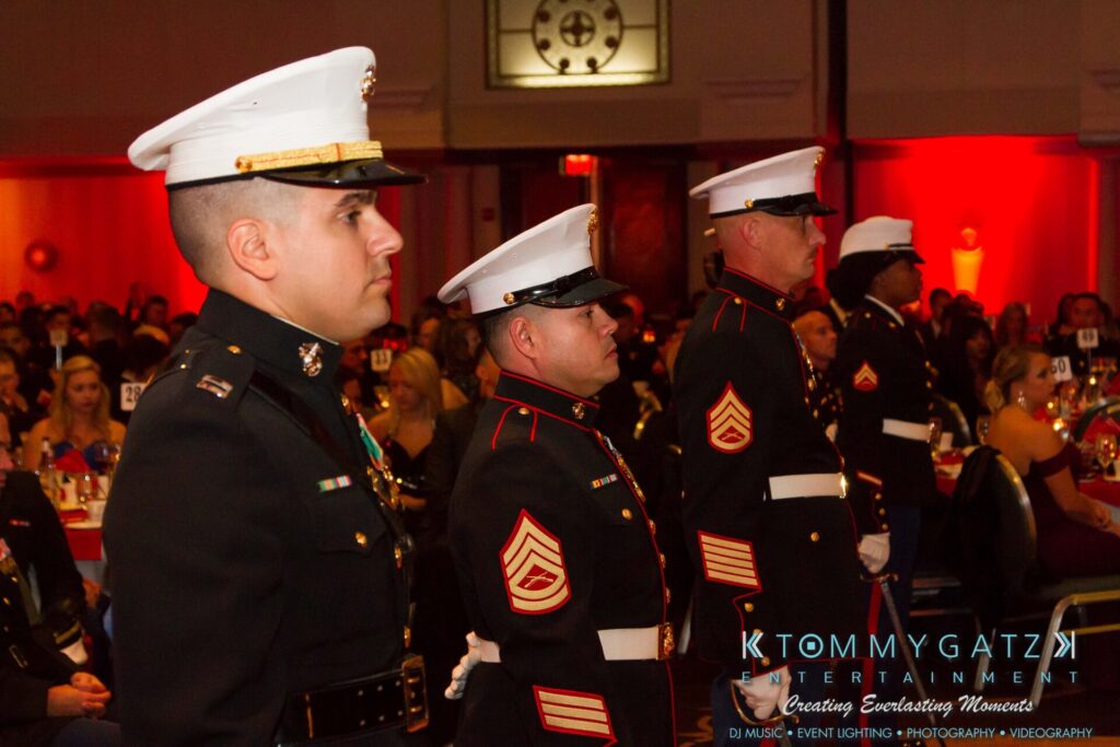 Marine-Lo-Res-30 - TOMMY GATZ ENTERTAINMENT A group of Marines in dress uniforms stand in formation at a formal event with a decorated hall and seated guests in the background.