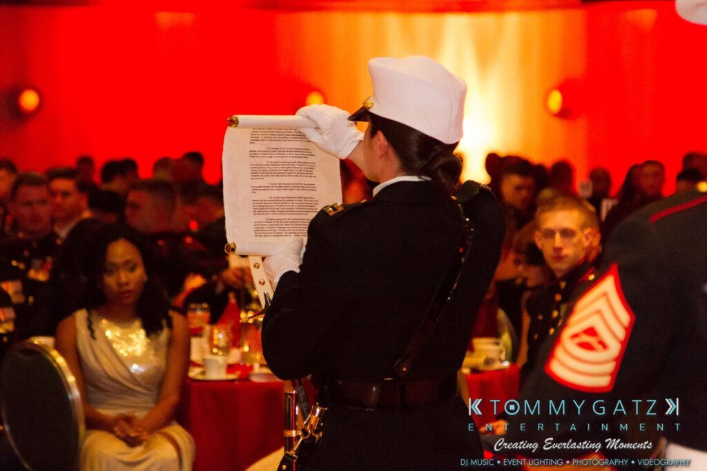 Marine-Lo-Res-32 - TOMMY GATZ ENTERTAINMENT A Marine reads from a document at a formal event, with people seated at tables in the background.