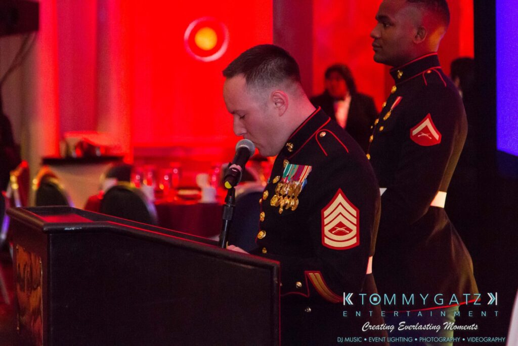 Marine-Lo-Res-33 - TOMMY GATZ ENTERTAINMENT A Marine in dress uniform speaks at a podium while another Marine stands nearby. The room is decorated with red lighting.