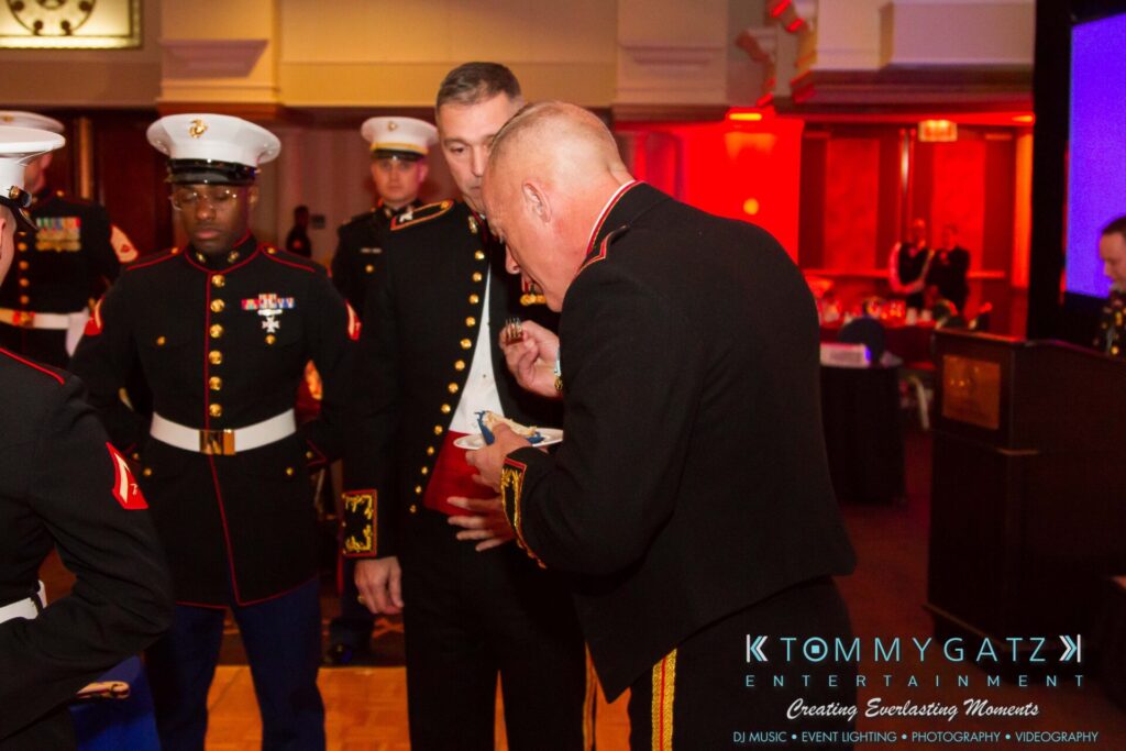 Marine-Lo-Res-37 - TOMMY GATZ ENTERTAINMENT A group of uniformed military personnel at an indoor event with red and blue lighting. A man in the foreground is writing in a notebook.