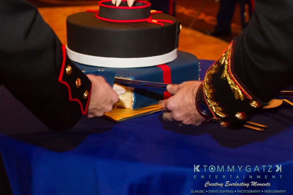 Marine-Lo-Res-39 - TOMMY GATZ ENTERTAINMENT Two uniformed individuals cut a blue and red cake with a ceremonial sword on a blue tablecloth, featuring a logo for Tommy Gatz Entertainment in the bottom right corner.