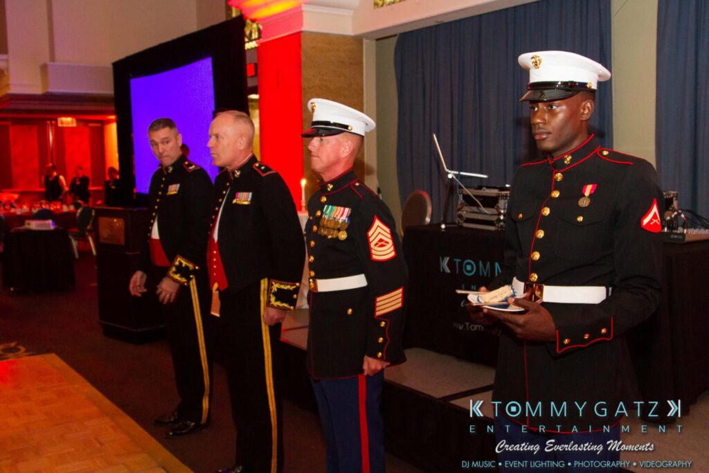 Marine-Lo-Res-42 - TOMMY GATZ ENTERTAINMENT Four men in military dress uniforms stand on stage; one holds an item. A DJ booth is visible in the background with event lighting.