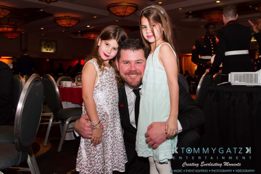 Marine-Lo-Res-5 - TOMMY GATZ ENTERTAINMENT A man in a suit kneels, smiling, with two young girls in dresses beside him at an indoor event.