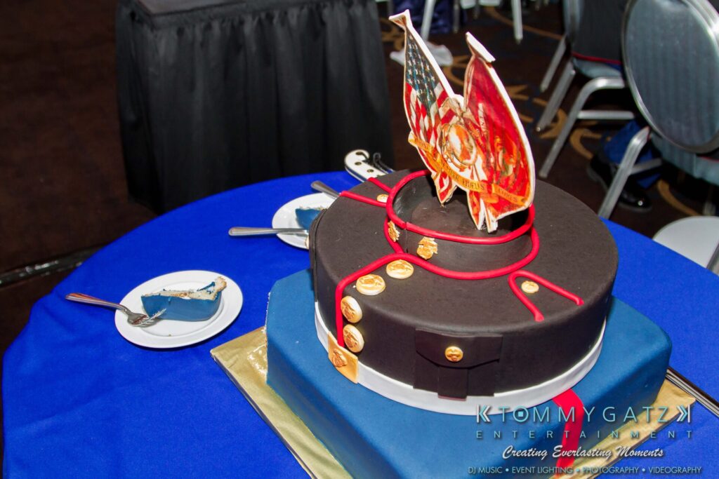Marine-Lo-Res-50 - TOMMY GATZ ENTERTAINMENT A two-tiered cake decorated in a military theme with emblem toppers and gold buttons. A slice is cut on a plate nearby on a blue tablecloth.