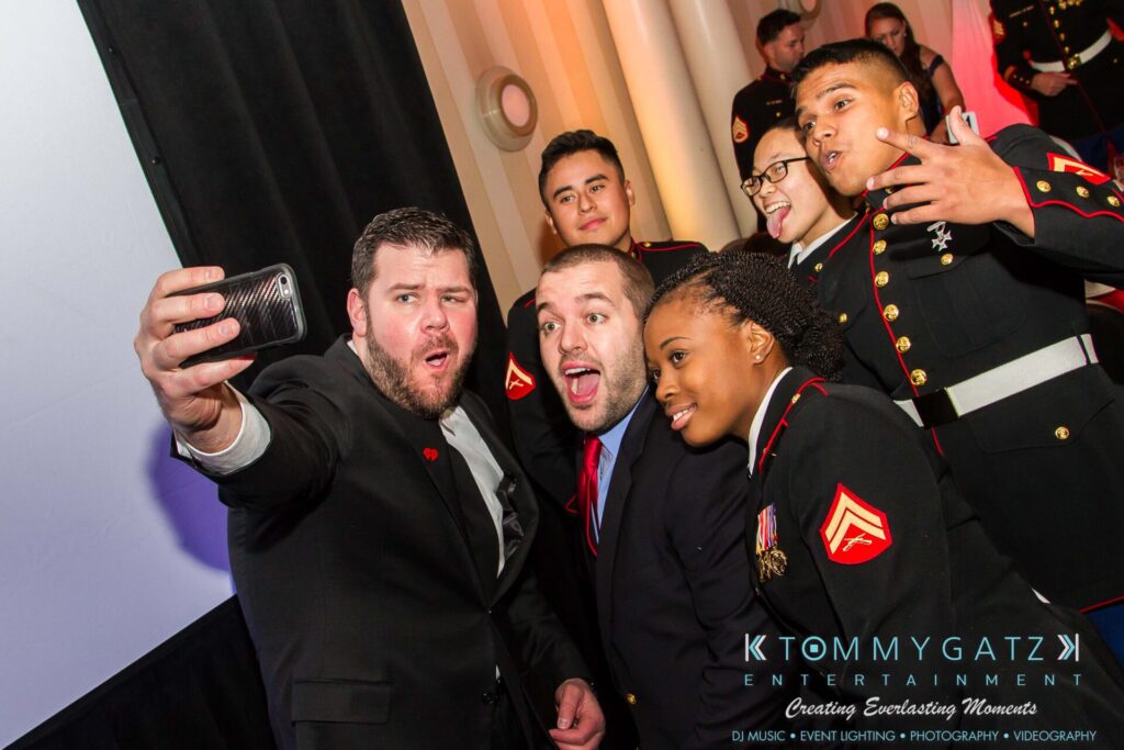Marine-Lo-Res-56 - TOMMY GATZ ENTERTAINMENT A group of people, some in military uniforms, take a selfie at an indoor event.