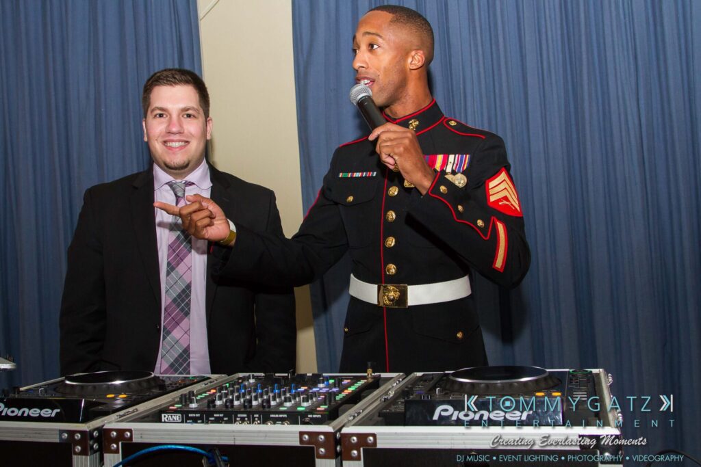 Marine-Lo-Res-57 - TOMMY GATZ ENTERTAINMENT A Marine in dress uniform speaks into a microphone while standing beside a man in a suit. They are in front of DJ equipment with a blue curtain in the background.