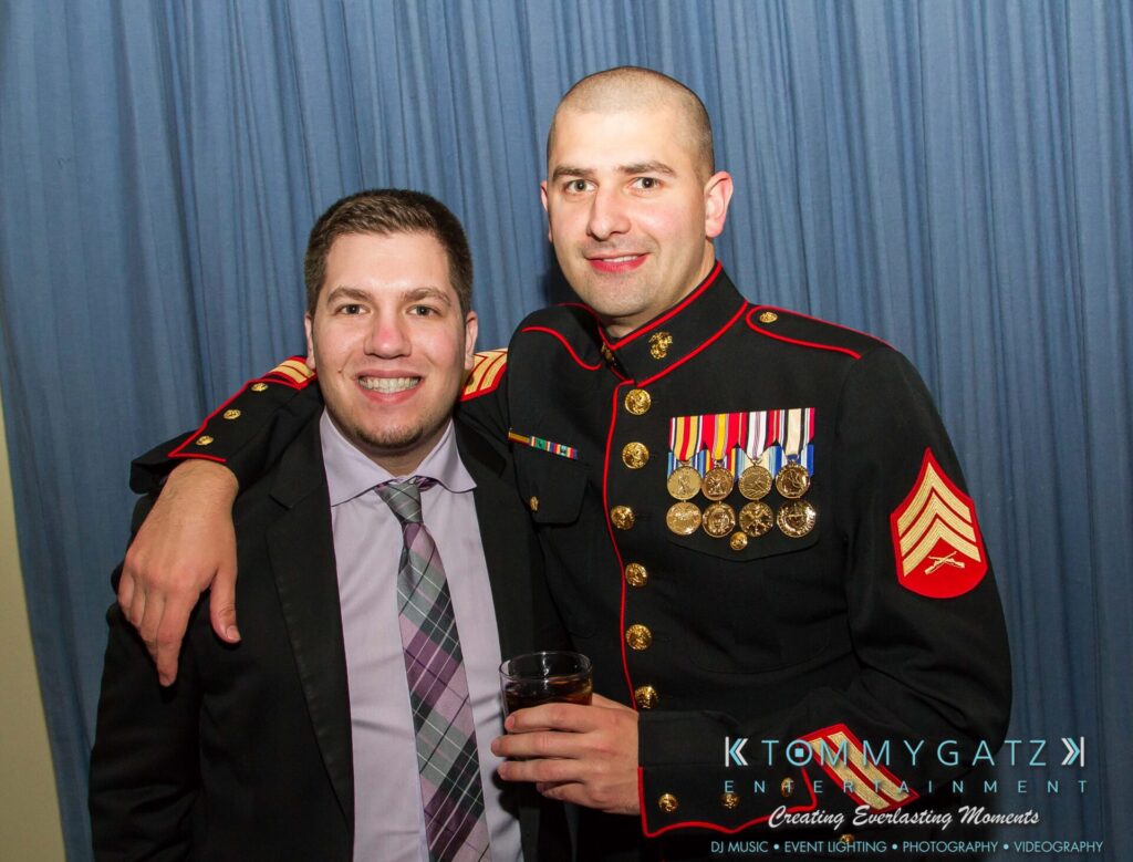 Marine-Lo-Res-66 - TOMMY GATZ ENTERTAINMENT Two men pose together; one in a suit and tie, the other in a military dress uniform with medals. Blue curtain backdrop.
