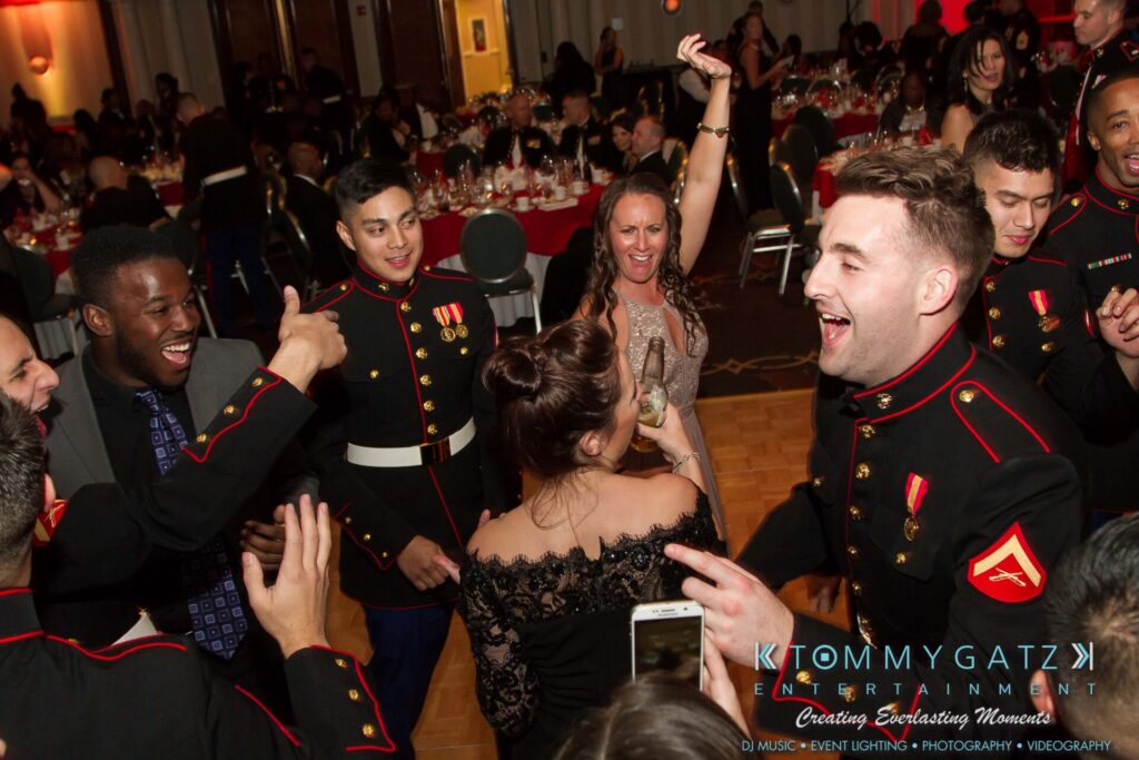 Marine-Lo-Res-73 - TOMMY GATZ ENTERTAINMENT Group of people in military dress uniforms and evening attire, laughing and dancing at an indoor event; tables set with red decor in the background.