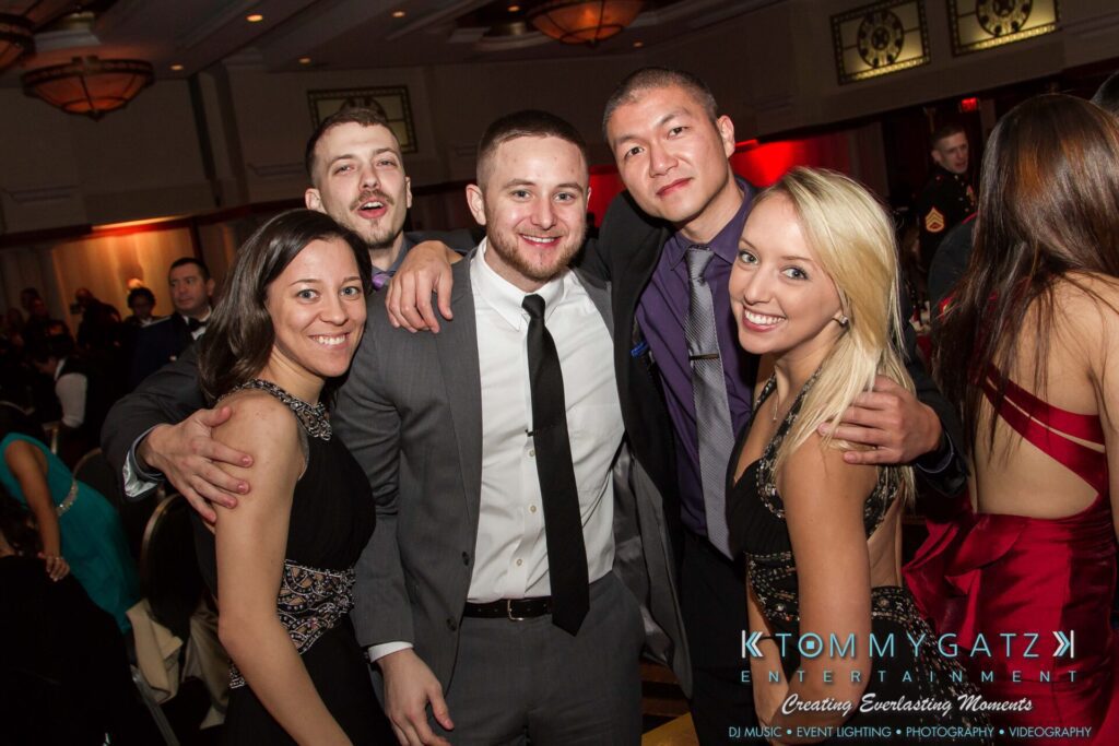 Marine-Lo-Res-84 - TOMMY GATZ ENTERTAINMENT A group of five people in formal attire pose together at an indoor event with others visible in the background.
