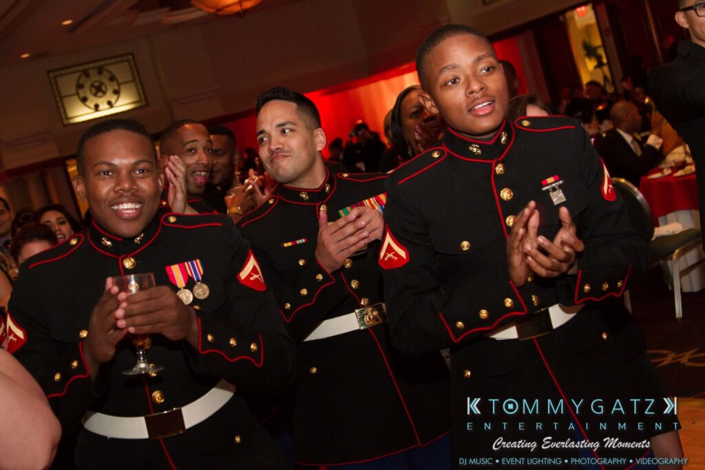 Marine-Lo-Res-97 - TOMMY GATZ ENTERTAINMENT Group of Marines in dress uniforms clapping and smiling at an event, with a logo at the bottom for "Tommy Gatz Entertainment.