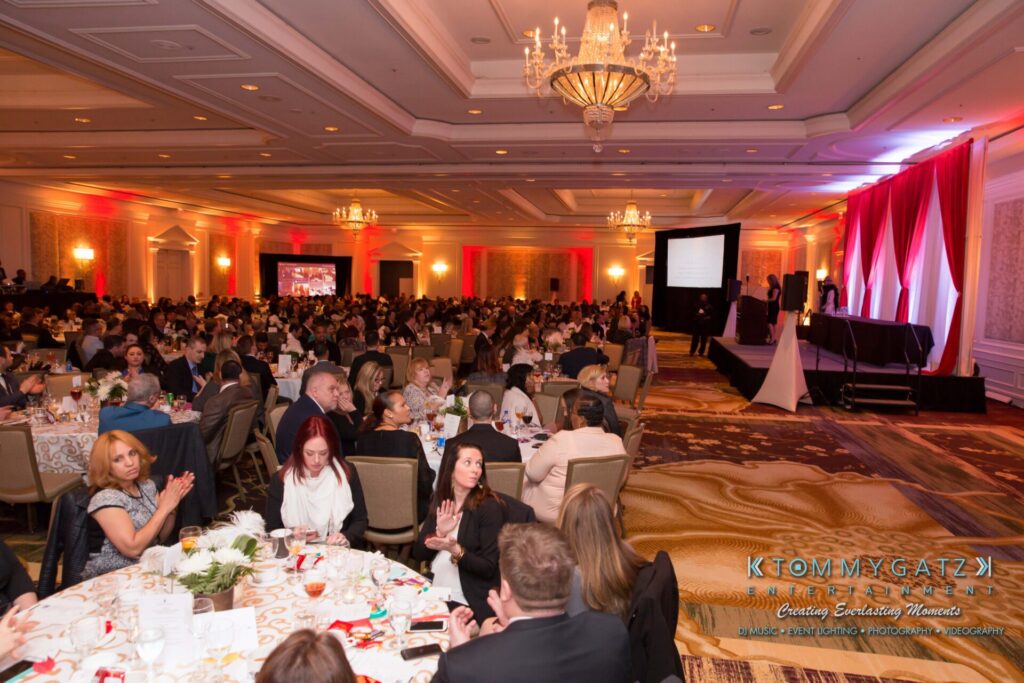 Pillar-Awards-32417-ADDITIONS-Blog-WM-11-1 - TOMMY GATZ ENTERTAINMENT Large banquet hall filled with people seated at tables, decorated with red and white drapes, and a stage with a projection screen.