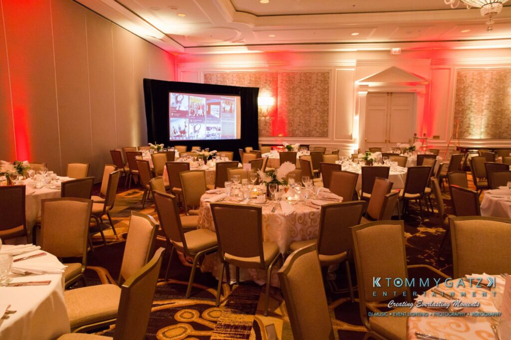 Pillar-Awards-32417-ADDITIONS-Blog-WM-2 - TOMMY GATZ ENTERTAINMENT A hotel banquet room is set up for an event with round tables, beige chairs, and a projection screen displaying images. The room is illuminated with warm red lighting accents.