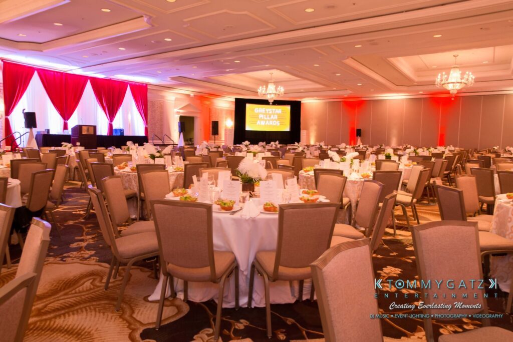 Pillar-Awards-32417-ADDITIONS-Blog-WM-4 - TOMMY GATZ ENTERTAINMENT A decorated banquet hall with round tables set for an event. A stage with red curtains and a lit sign is at the front.