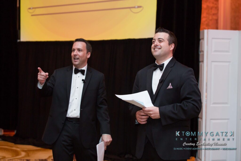 Pillar-Awards-32417-ADDITIONS-Blog-WM-6 - TOMMY GATZ ENTERTAINMENT Two men in tuxedos stand on stage, smiling and holding papers. One points while the other looks ahead.
