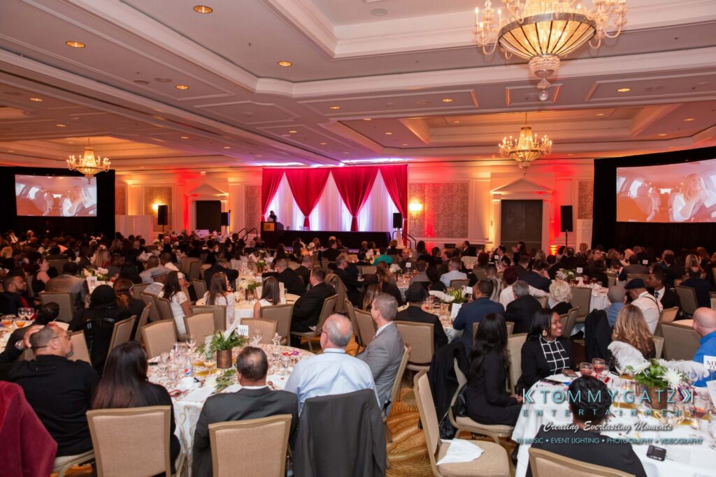 Pillar-Awards-32417-ADDITIONS-Blog-WM-8 - TOMMY GATZ ENTERTAINMENT A large audience at a formal event in a banquet hall with round tables, red lighting, and presentation screens.