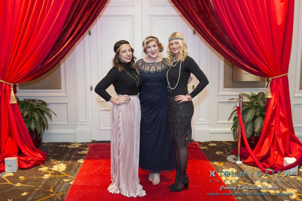 Pillar-Awards-Final-3-24-17-Blog-WM-11 - TOMMY GATZ ENTERTAINMENT Three women pose on a red carpet, dressed in vintage-style outfits with headbands and pearl necklaces. Red curtains and a roped-off area are in the background.