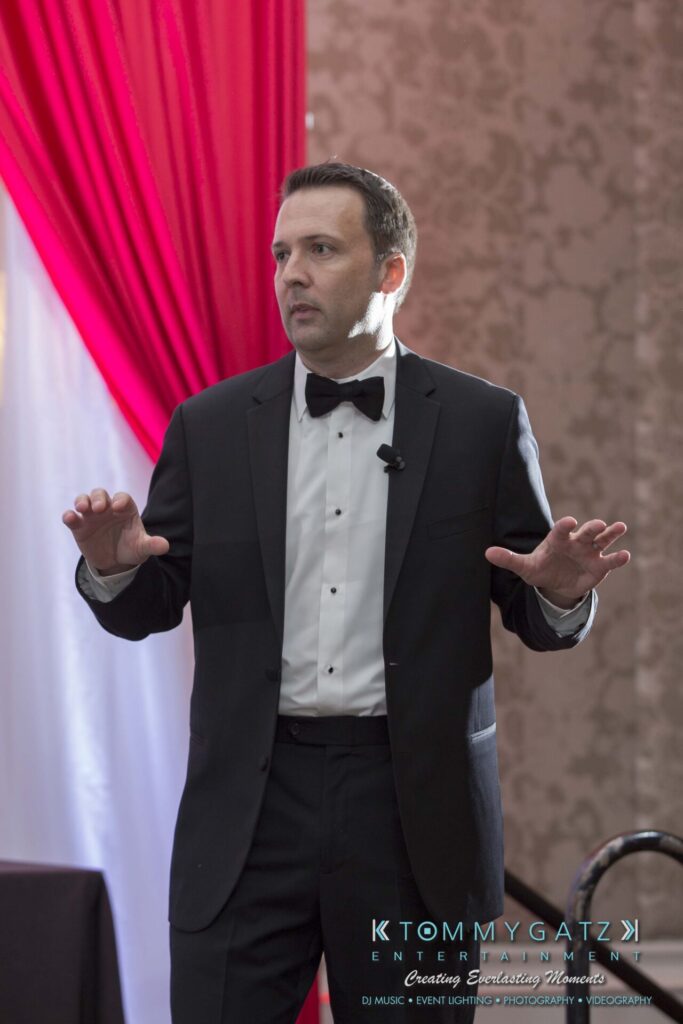 Pillar-Awards-Final-3-24-17-Blog-WM-111 - TOMMY GATZ ENTERTAINMENT A man in a tuxedo stands in front of red and white drapes, gesturing with both hands.