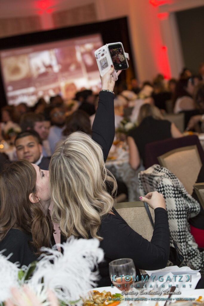 Pillar-Awards-Final-3-24-17-Blog-WM-113 - TOMMY GATZ ENTERTAINMENT Two people seated at a table taking a selfie with a smartphone at an indoor event. The room has a crowd and red lighting.