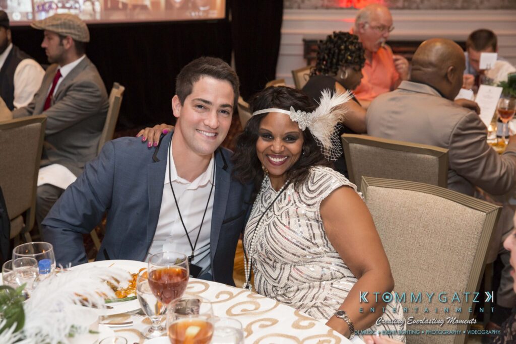 Pillar-Awards-Final-3-24-17-Blog-WM-119 - TOMMY GATZ ENTERTAINMENT Two people sitting at a banquet table, smiling at the camera. The woman is wearing a feathered headpiece, and others are seated in the background.