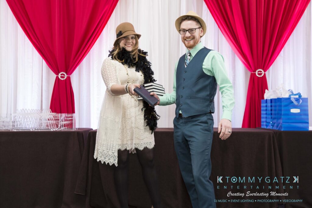 Pillar-Awards-Final-3-24-17-Blog-WM-139 - TOMMY GATZ ENTERTAINMENT Two people stand in front of a table with red and white drapes. One hands a small stack of books to the other. They are smiling and dressed formally.